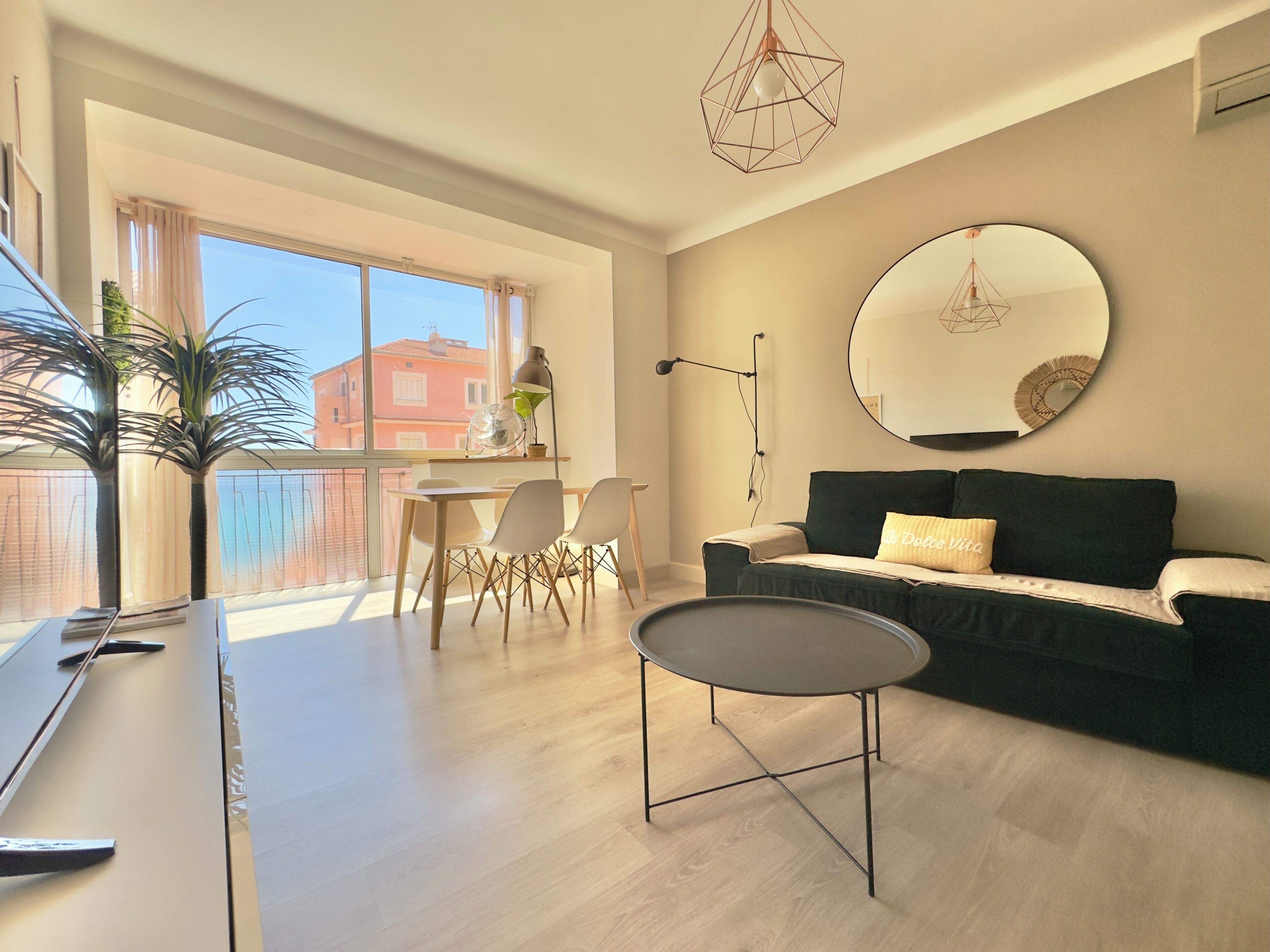 A modern living room with a sofa, a round mirror, a dining table with chairs, and a large window with curtains.
