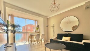 Ein modernes Wohnzimmer mit einer Couch, einem runden Spiegel, einem Esstisch mit Stühlen und einem großen Fenster mit Vorhängen.