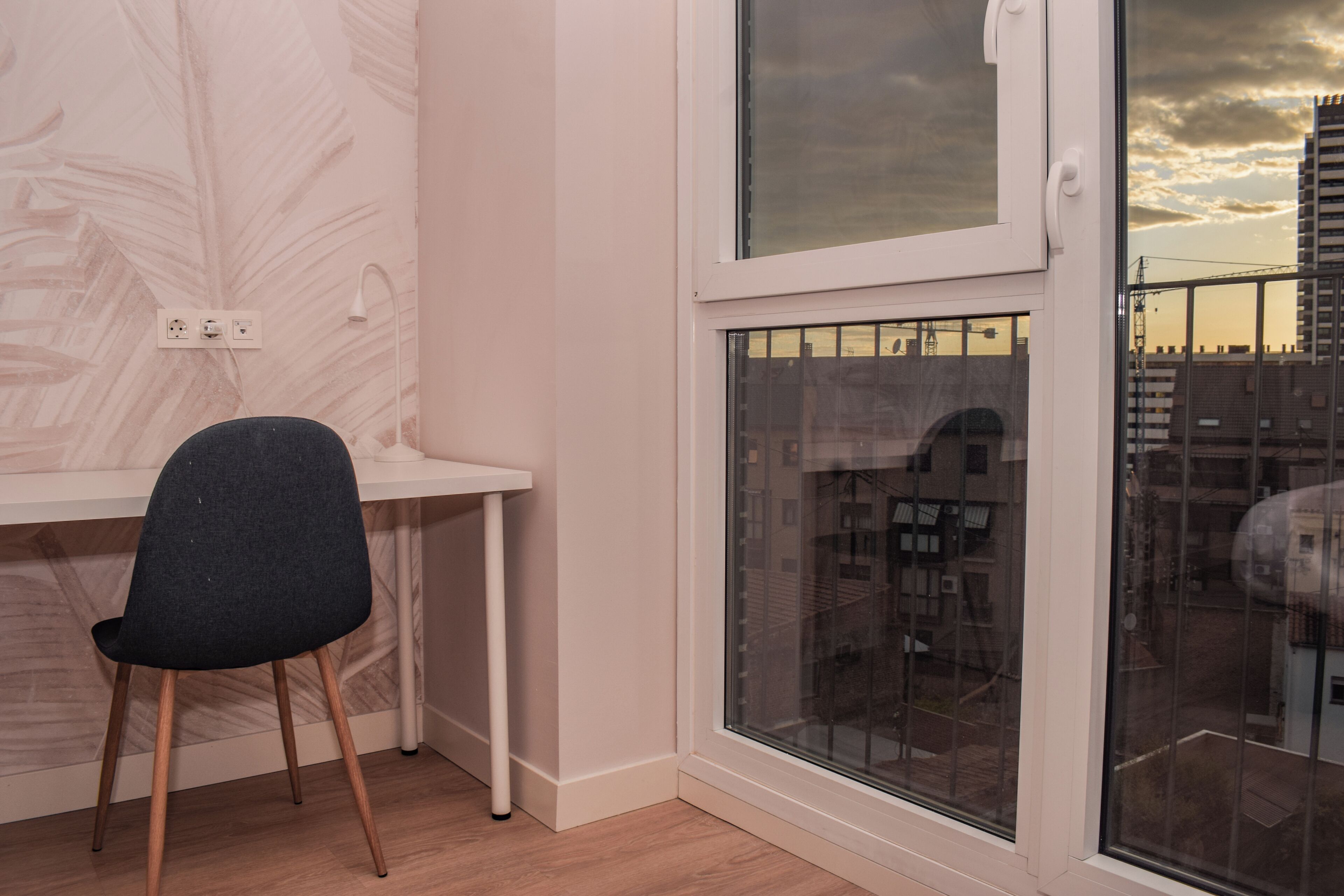 Una habitación con escritorio, silla, ventana con vista a edificios y toma de corriente en la pared.