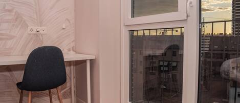 Una habitación con escritorio, silla, ventana con vista a edificios y toma de corriente en la pared.