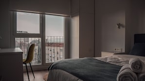 A modern hotel room with a large window, a bed with rolled towels, a desk, and a chair.
