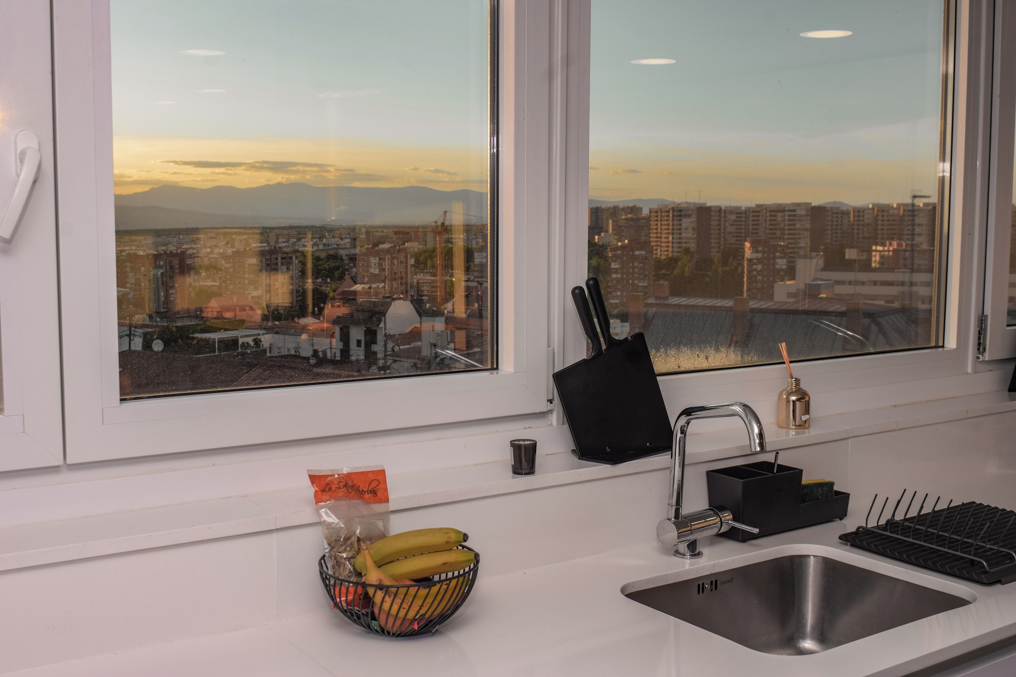 A kitchen with a large window, a sink, a faucet, a knife block, and a fruit bowl.