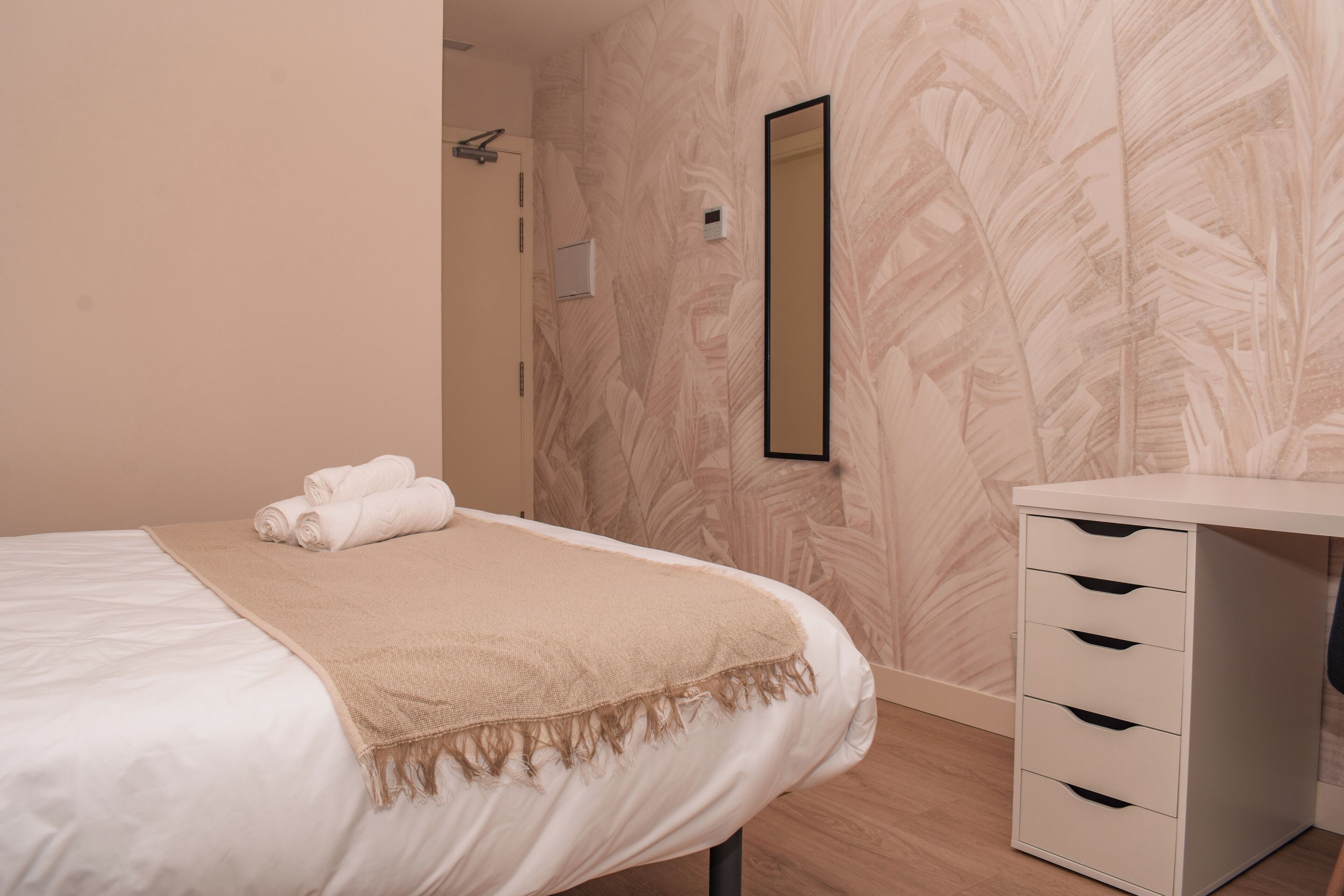 Una cama bien hecha con una manta color beige, una mesita de noche blanca con cajones, y una pared con diseño de hojas.