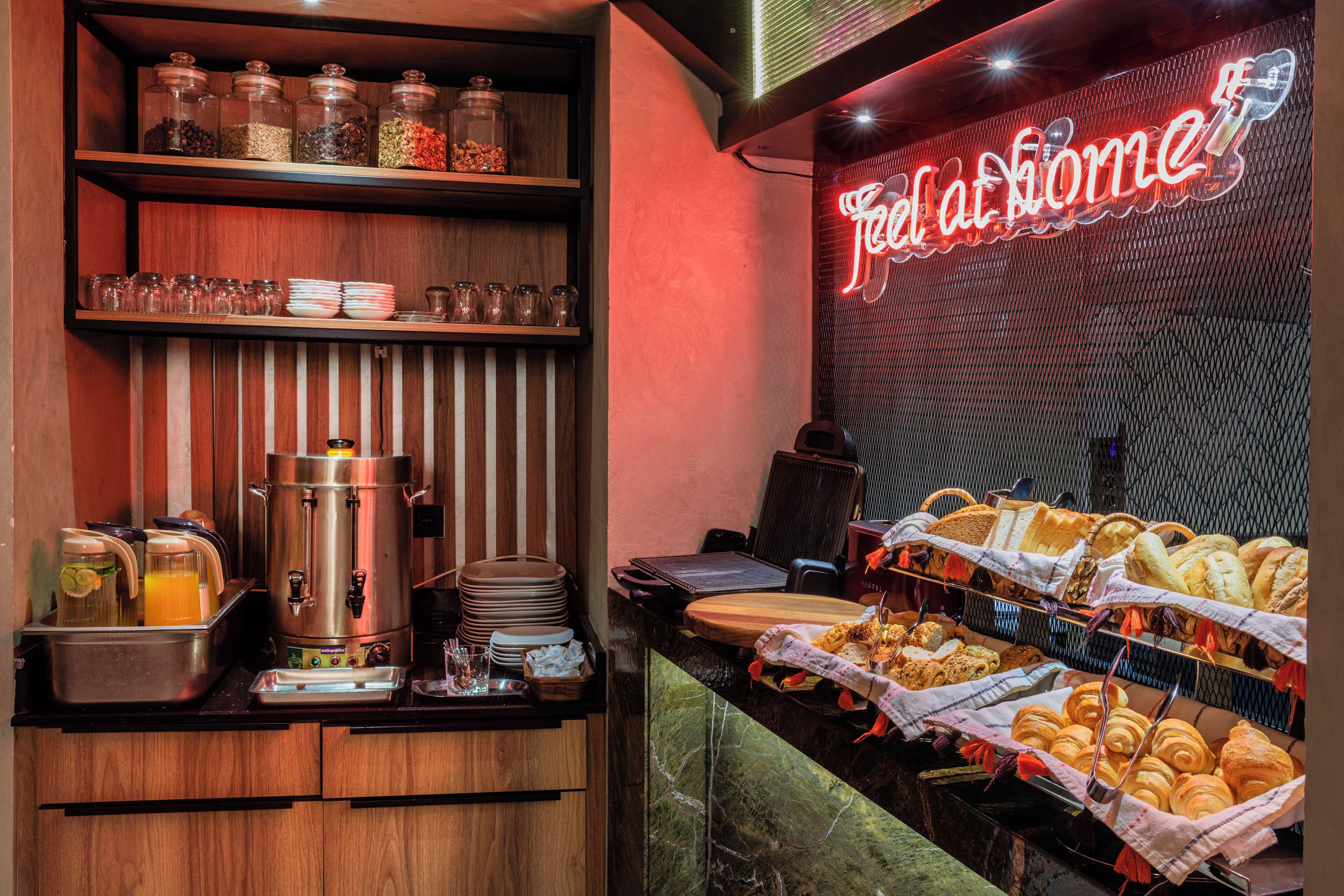A breakfast buffet with various food items, a coffee machine, and a neon sign reading 'Feel at home'.