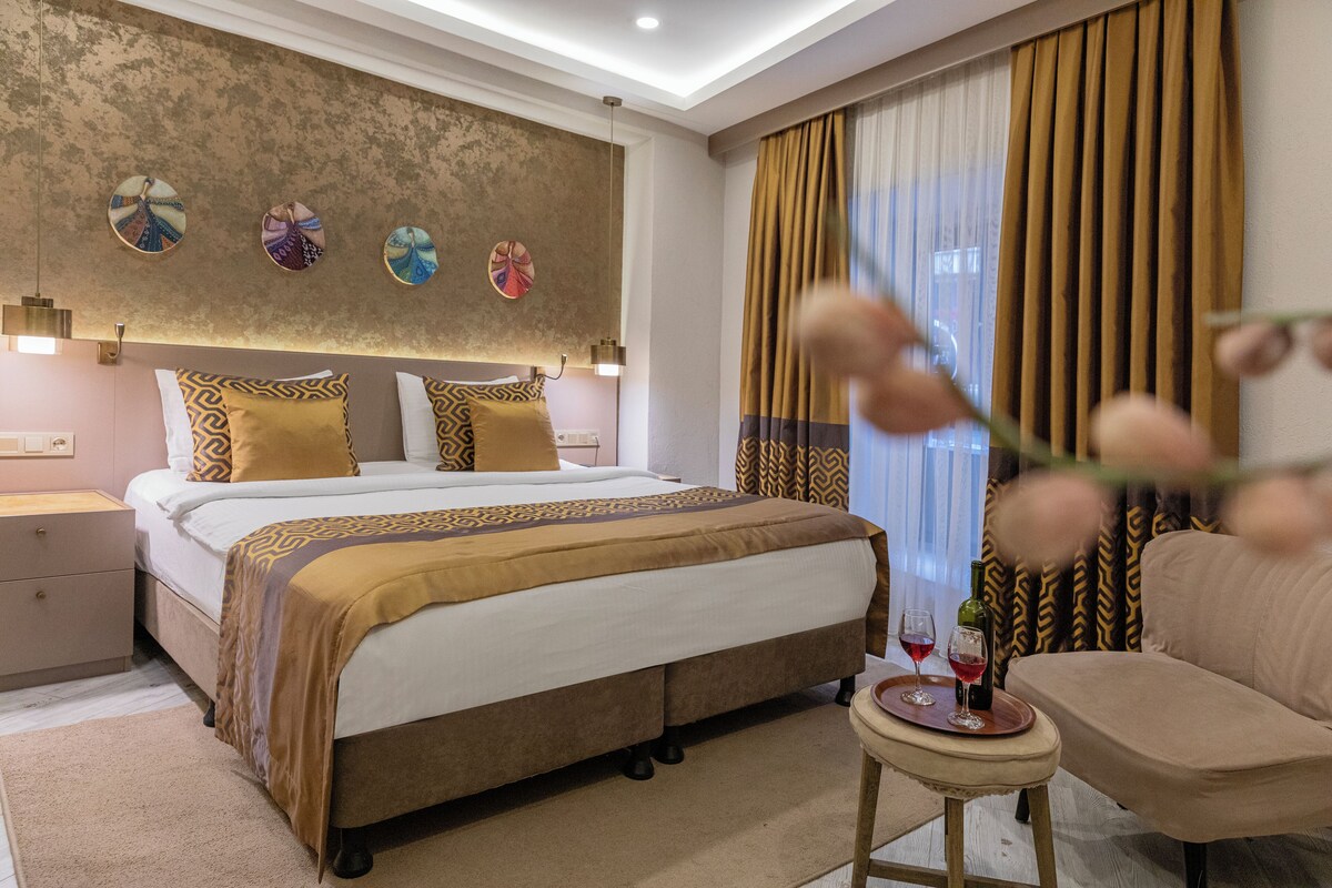 Ein modernes Hotelzimmer mit einem großen Bett, einem Nachttisch, einem kleinen runden Tisch mit Weingläsern und einer Flasche sowie einem Sofa.
