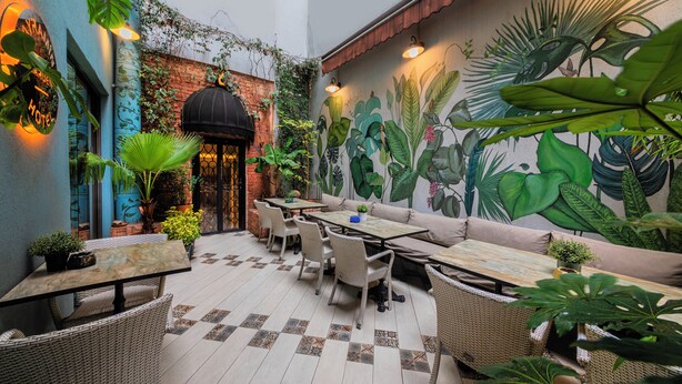 Un patio al aire libre con mobiliario de mimbre, un mural de plantas tropicales y una pared de ladrillo.