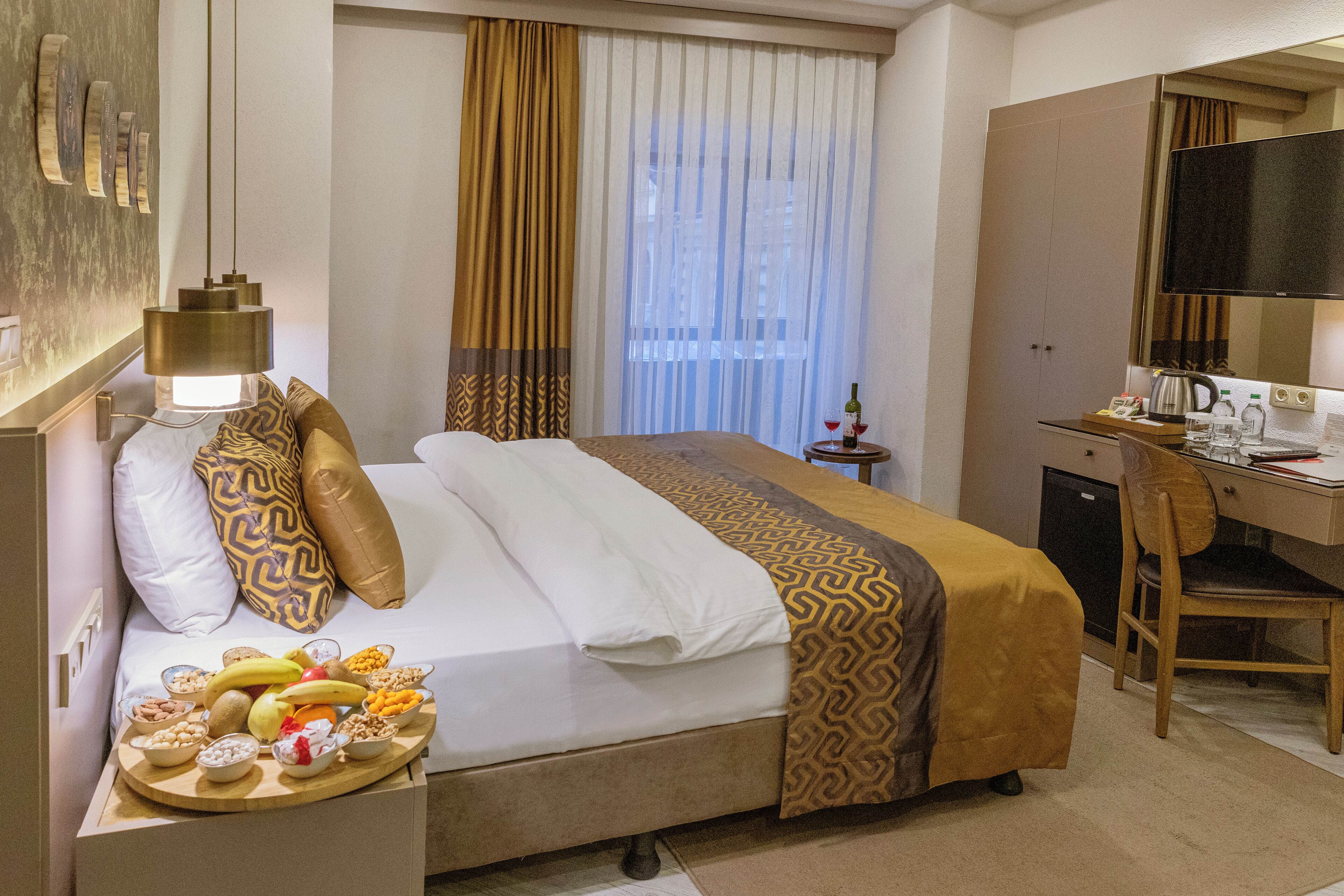 A hotel room with a bed, a desk, a chair, a television, a small table with snacks, and a window with curtains.
