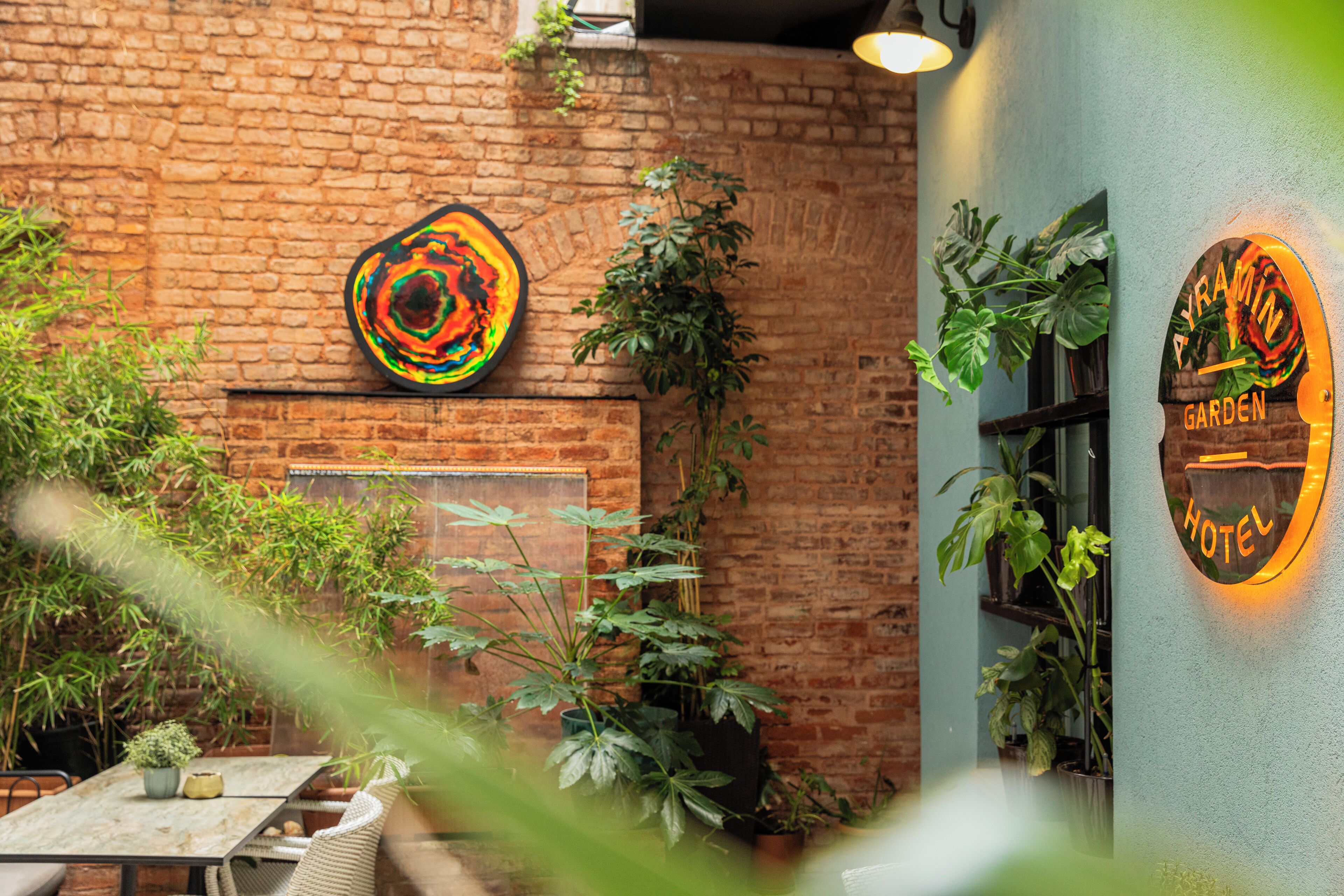 A brick wall with a colorful abstract painting, a sign for 'Ayramin Garden Hotel,' and various potted plants.
