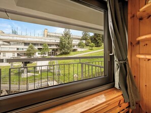 Une fenêtre donnant sur un ensemble d’immeubles, une zone herbeuse et une rambarde en métal.