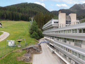 A modern hotel with multiple floors, a parking area, and a view of a forested mountain. - Alpen Nook - Sci e Natura by Interhome (Marilleva 1400)