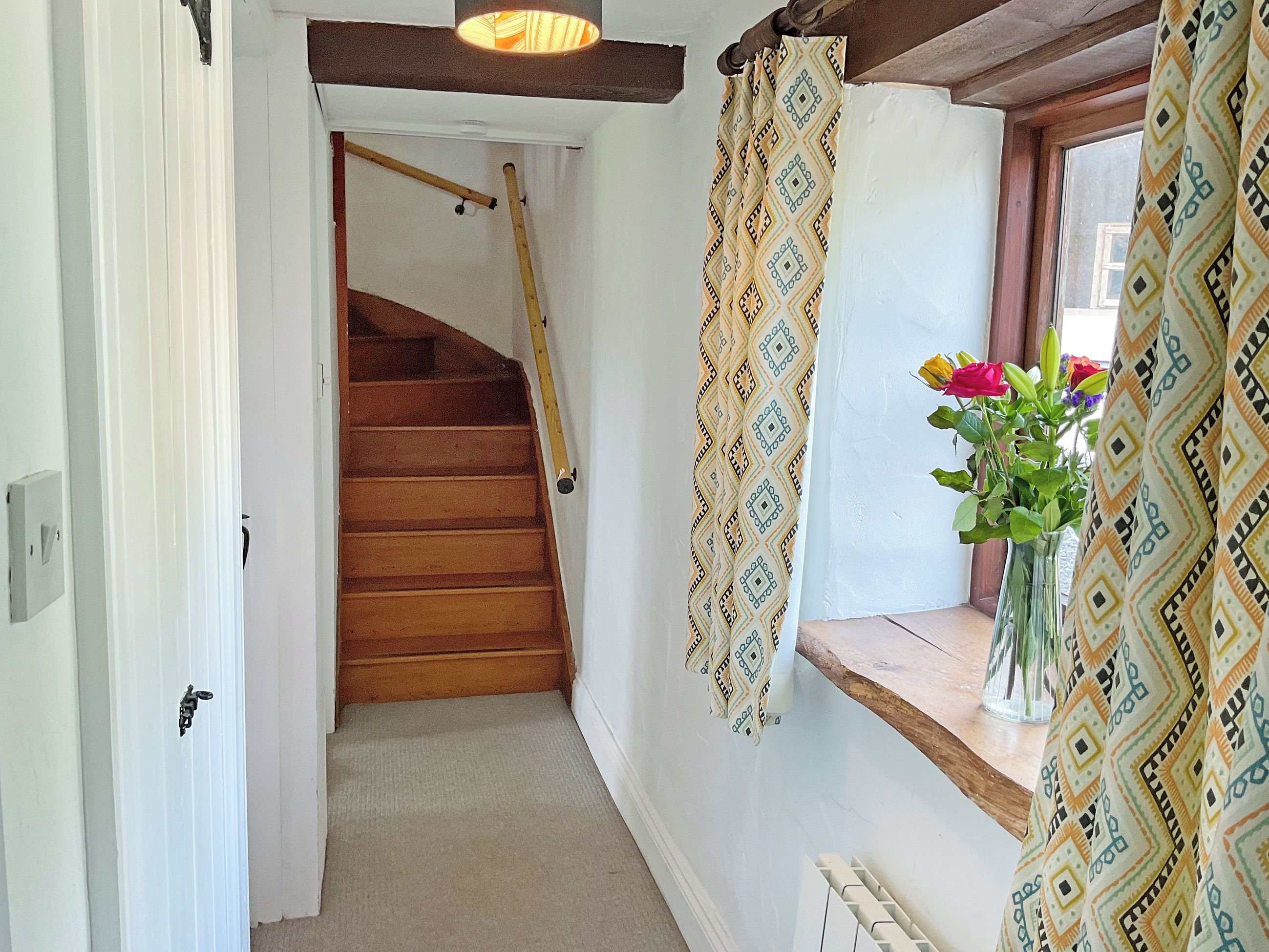Eine Treppe führt zu einer Zwischenebene, ein Fenster mit gemusterten Vorhängen und ein Blumenvasen auf einem Fensterbrett.