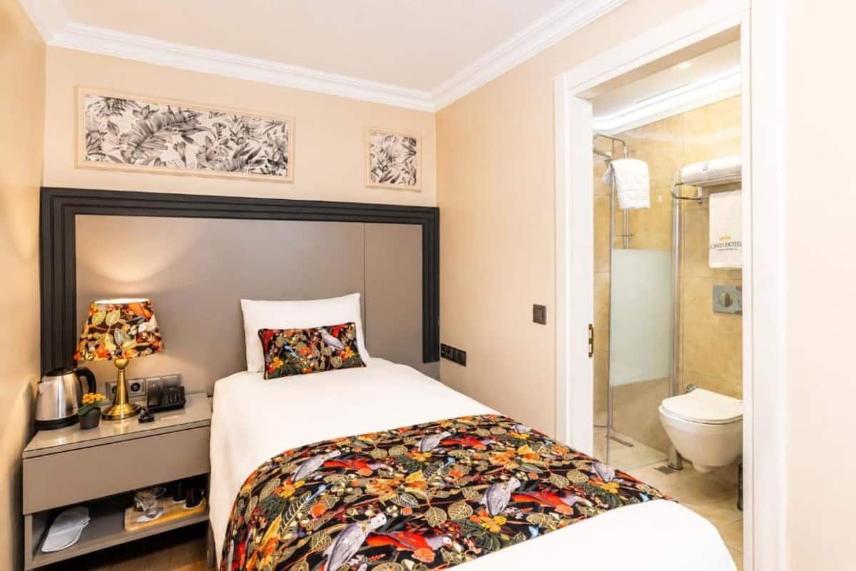A hotel room with a bed, a bedside table with a lamp, a coffee maker, and a framed floral artwork on the wall.