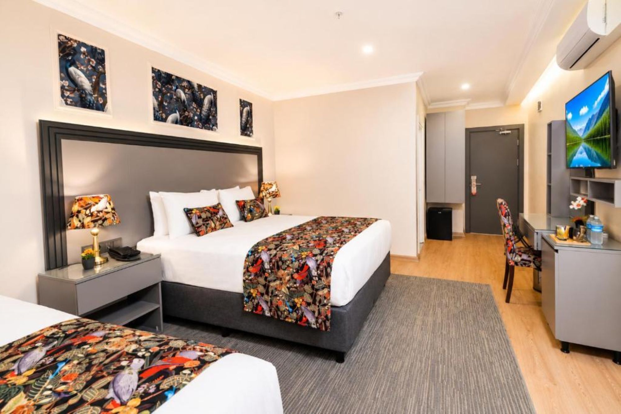 A hotel room with a bed, bedside tables, a desk, a chair, a TV, and artwork on the walls.