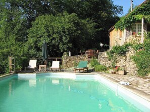 Une piscine avec des chaises longues, un mur en pierre et un bâtiment en bois surmonté d’un toit en pierre.