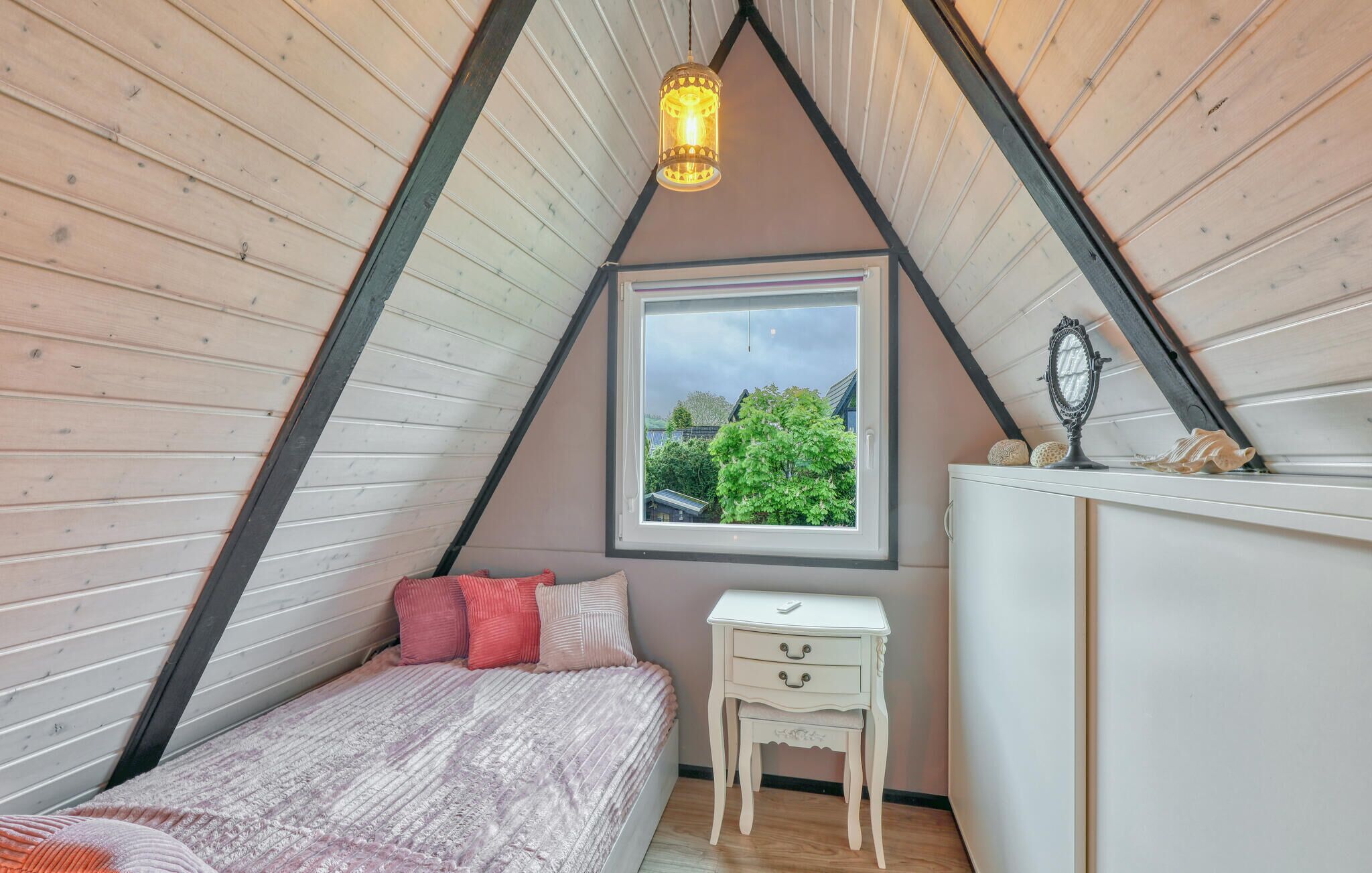 A bedroom with a sloped ceiling, a window with a view of trees, a bed with pink bedding, a small bedside table, and a dresser.
