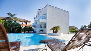 Uma piscina com espreguiçadeiras, um edifício branco com varandas e vegetação ao redor.