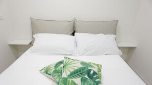 Uma cama com lençóis brancos, duas almofadas e um cobertor estampado com folhagens tropicais.