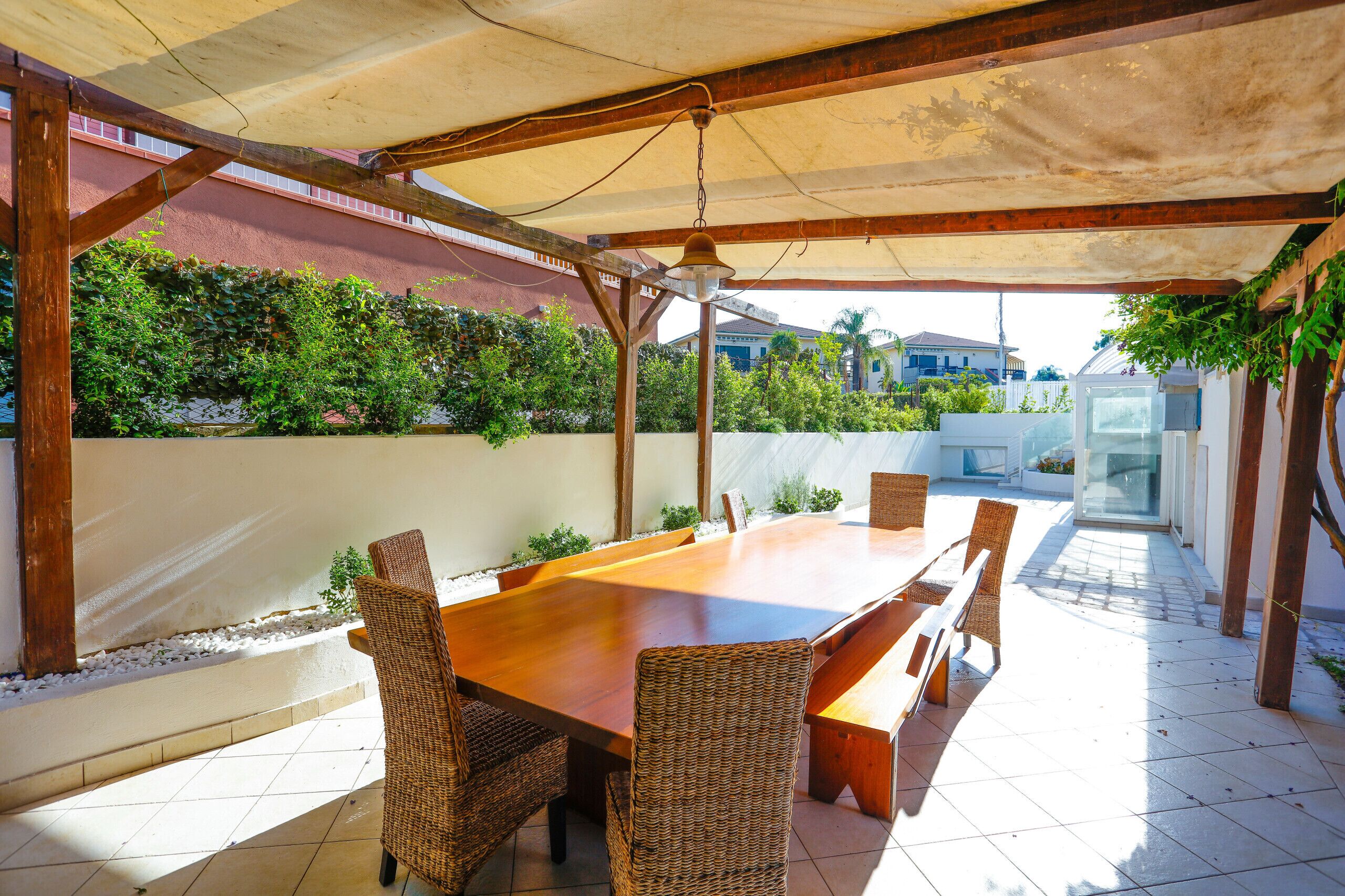 An outdoor patio with a wooden table and wicker chairs, surrounded by a covered area with a view of a residential neighborhood.