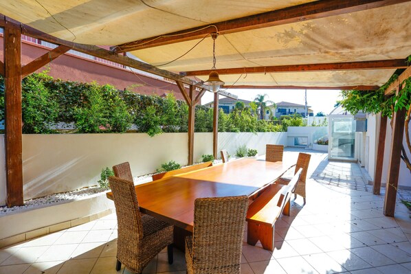 Eine Terrasse im Freien mit einem Holztisch und Korbstühlen, umgeben von einem überdachten Bereich mit Blick auf eine Wohngegend.