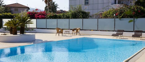 A swimming pool with lounge chairs, a white wall, and a building in the background.
