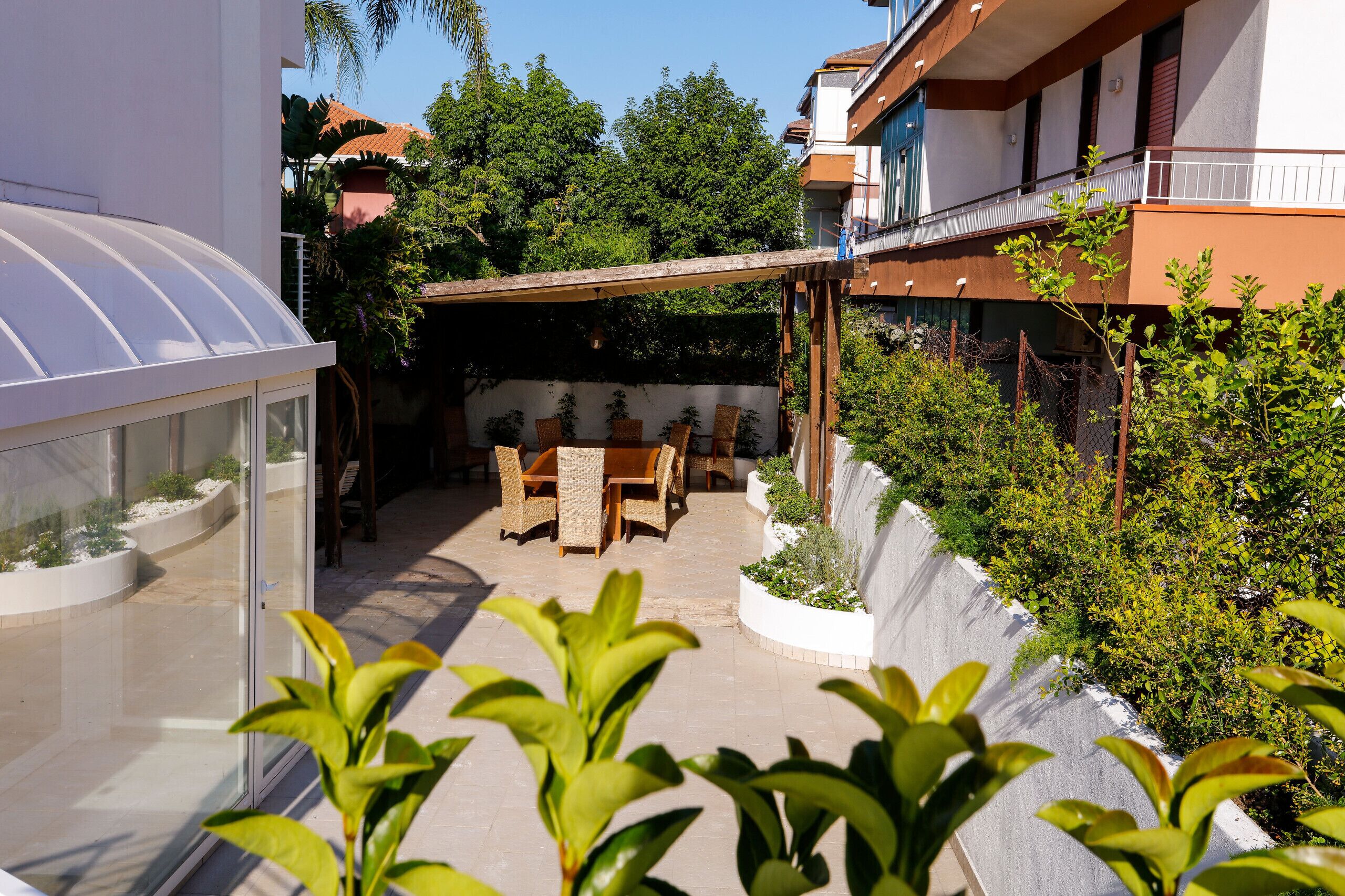A covered patio with a table and chairs, surrounded by plants and a glass greenhouse structure.