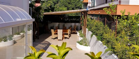 A covered patio with a table and chairs, surrounded by plants and a glass greenhouse structure.