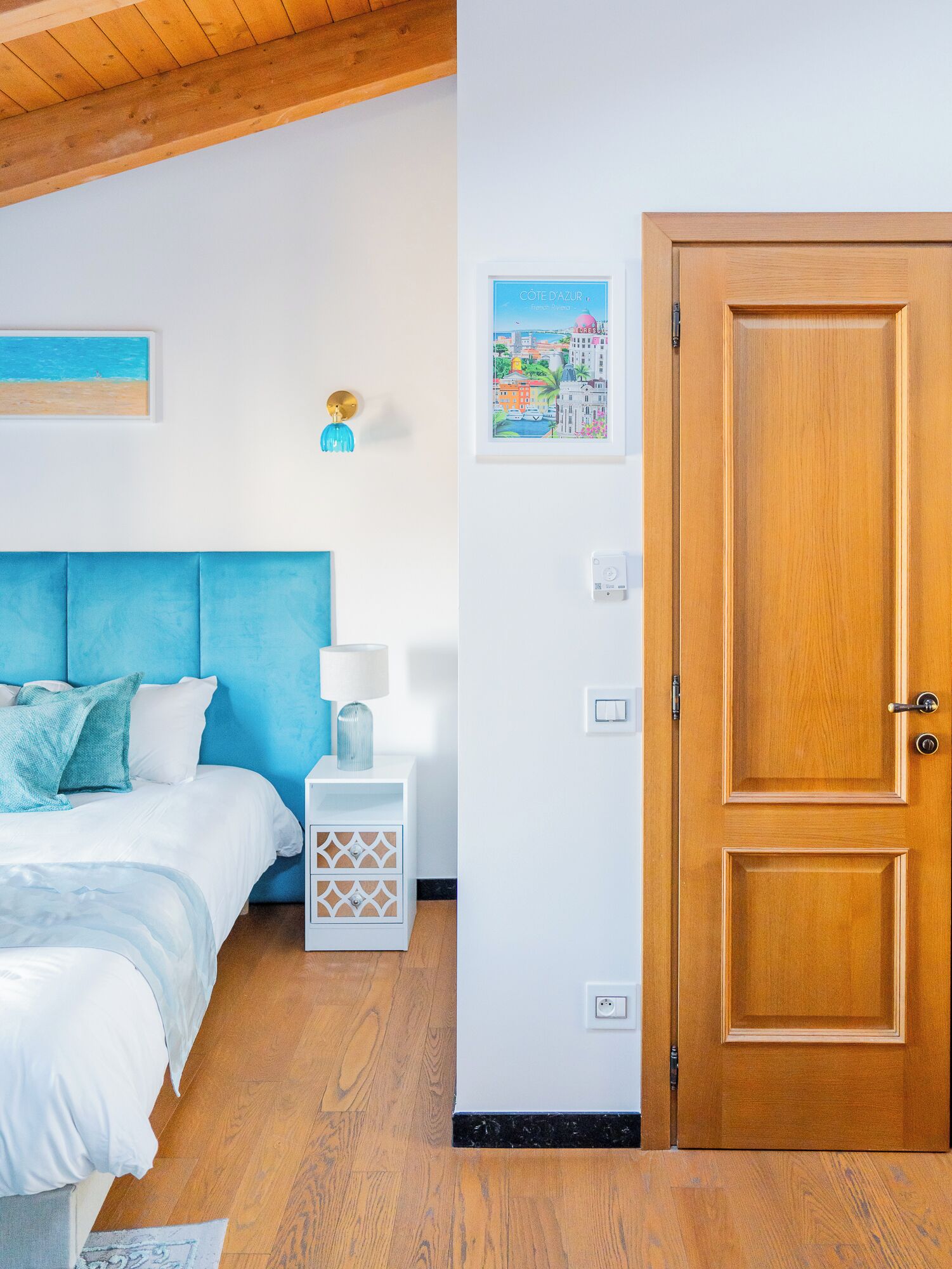 A bedroom with a bed, bedside table, lamp, wall art, and a wooden door.