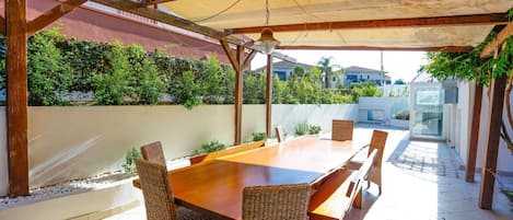 An outdoor patio with a wooden table and wicker chairs, surrounded by a covered area with a view of a residential neighborhood.