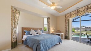 Une chambre à coucher avec un lit, un ventilateur de plafond, une table de chevet et une vue sur une piscine, visible à travers de grandes fenêtres.