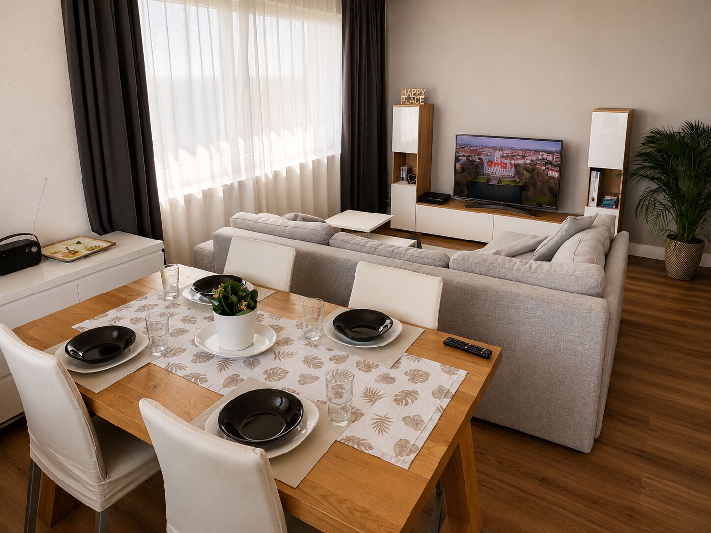 Un salon moderne avec un canapé, une table à manger dressée pour deux personnes, un téléviseur et une plante.
