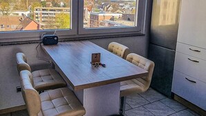 A compact kitchen with a dining table, chairs, a trash bin, and a view of the city through large windows.