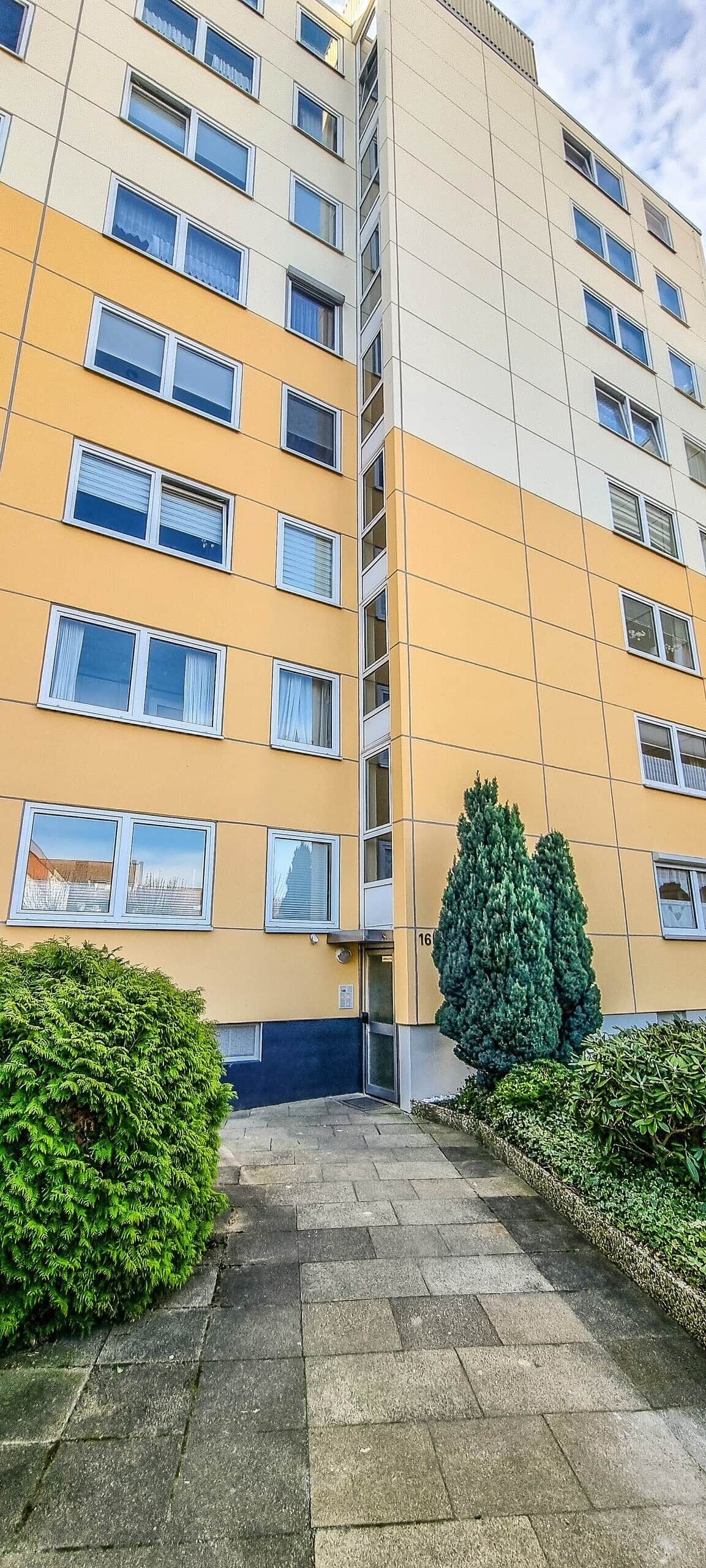 Un immeuble à plusieurs étages, doté d’une façade jaune, de nombreuses fenêtres et d’un espace d’entrée pavé agrémenté de verdure.