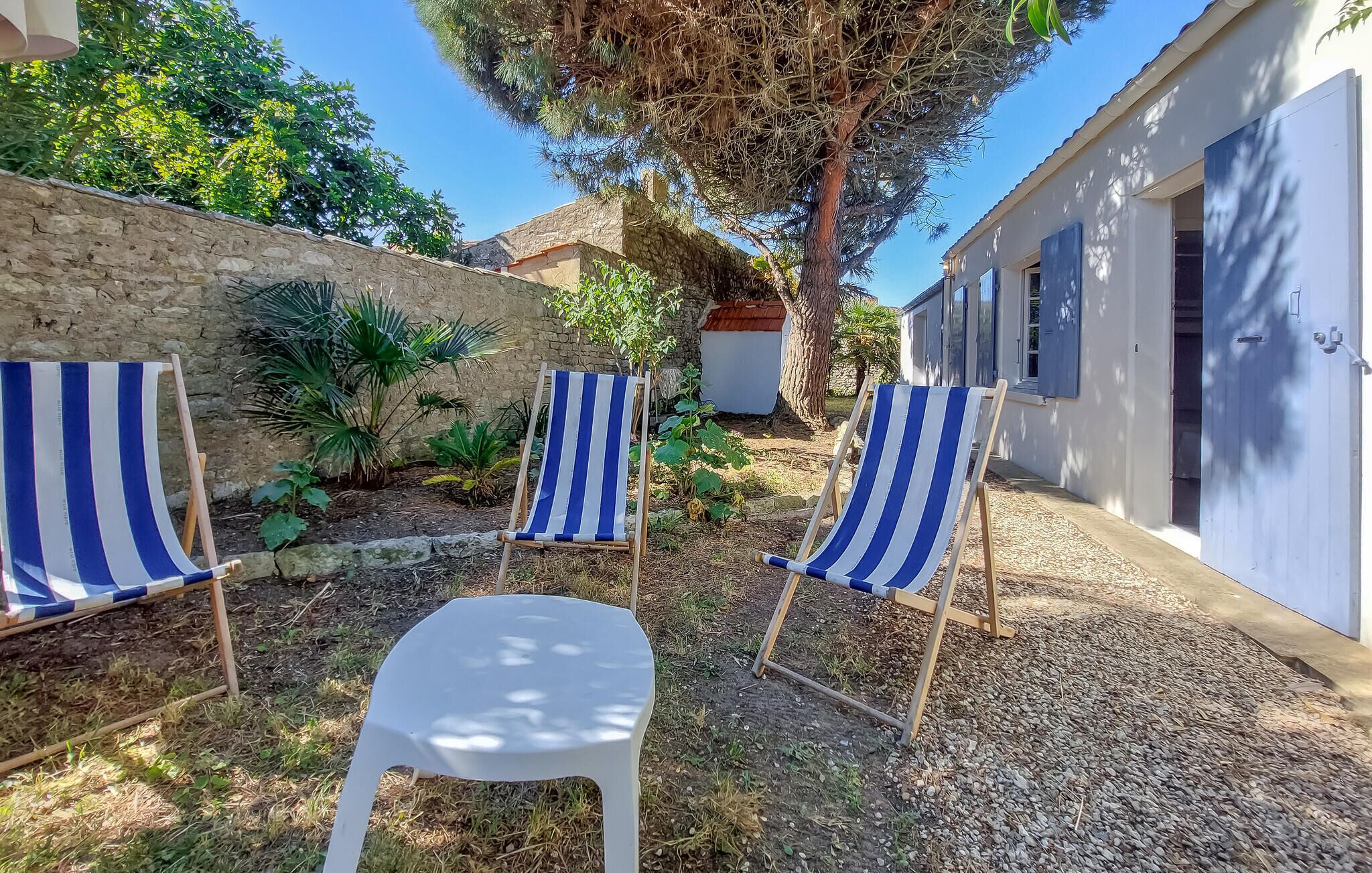 Trois chaises de plage à rayures et une chaise en plastique blanche, dans un espace jardin agrémenté d’un mur en pierre et d’un arbre.