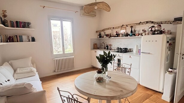 Een knus woongedeelte met een eettafel, stoelen, een bank, een keuken met een koelkast, en een raam dat voor natuurlijk licht zorgt.