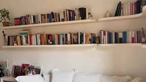 Een witte bank met een plaid, een witte bijzettafel met een vaas en boeken, en een boekenkast met verschillende boeken en decoratieve objecten.