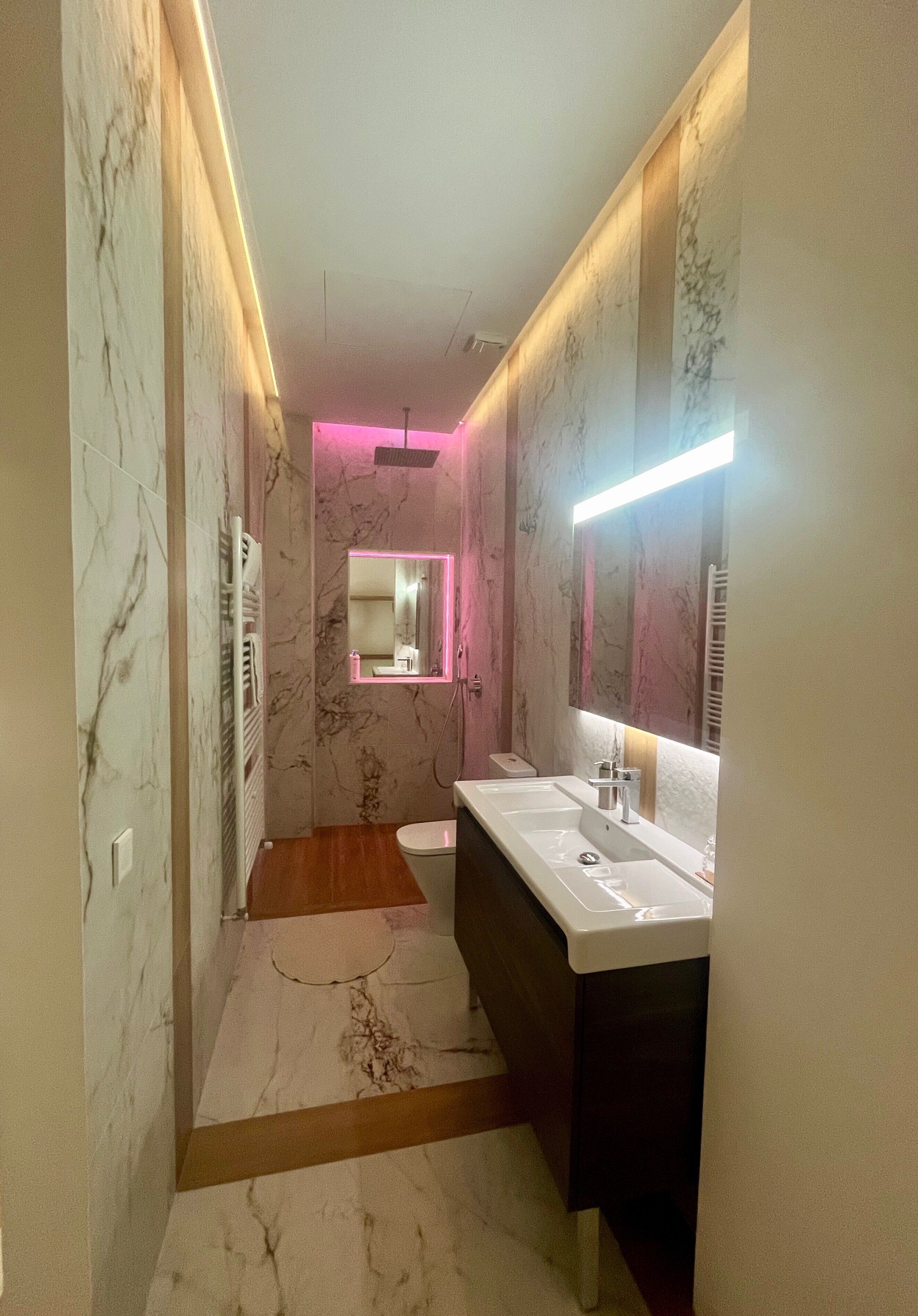 Un baño moderno con paredes de mármol, suelo de madera, un lavabo blanco y un espejo con luz de neón.