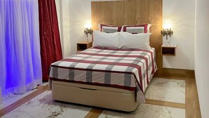 A bedroom with a bed, wooden ceiling beams, a ceiling fan, a nightstand with a lamp, and a marble-patterned floor.