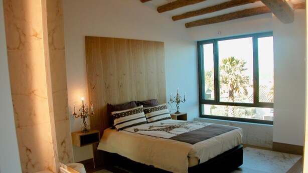 A bedroom with a bed, a nightstand, a candelabra, a window with a view, and a wooden ceiling.