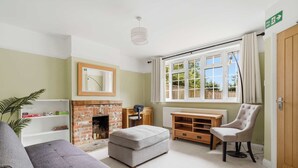A room with a fireplace, a sofa, an ottoman, a chair, a wooden cabinet, and a window with curtains.