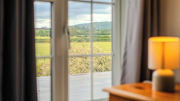 A window with a view of a green landscape, a wooden table with a lamp, and a curtain partially drawn.