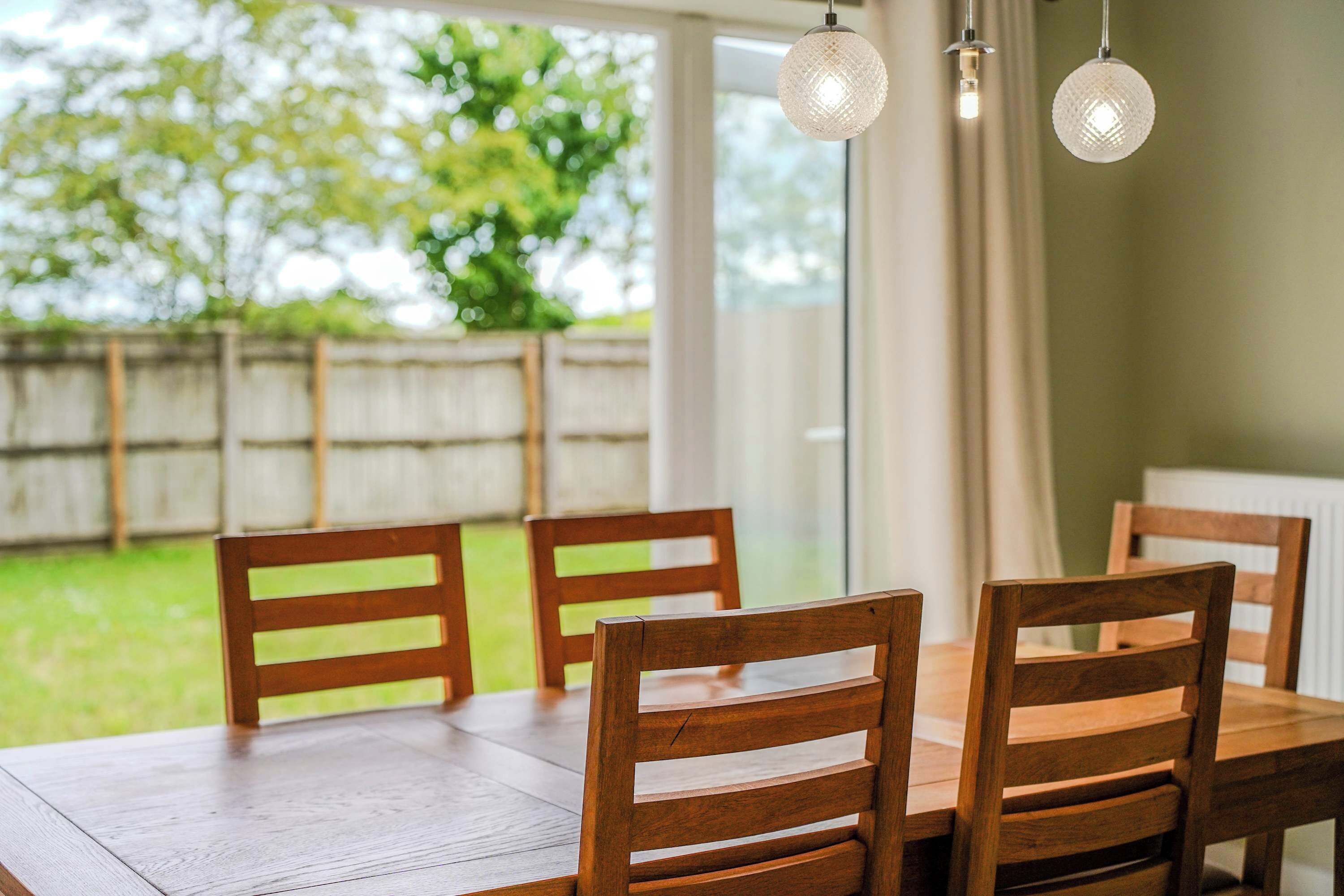 A wooden dining table with four chairs, a hanging light fixture, and a view of a green outdoor area through a window.
