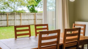 A wooden dining table with four chairs, a hanging light fixture, and a view of a green outdoor area through a window.