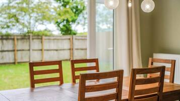 A wooden dining table with four chairs, a hanging light fixture, and a view of a green outdoor area through a window.