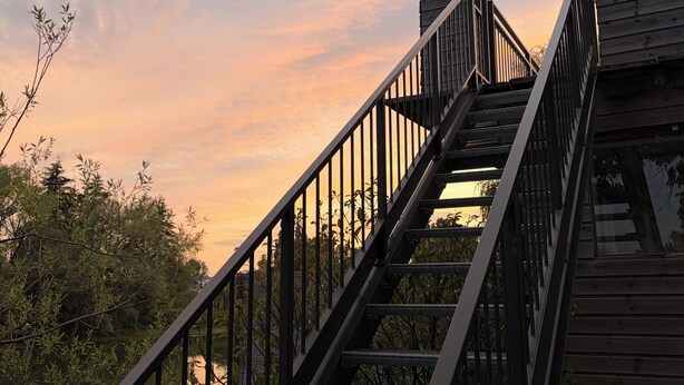 Een houten trap met metalen leuningen die naar een gebouw leidt, omringd door bomen en een zonsondergang.