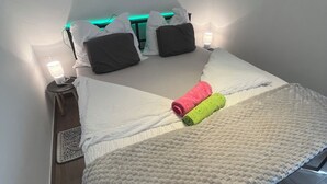 A neatly made bed with a white duvet, pillows, and a rolled towel. There are bedside lamps and a wall-mounted light. - Beautiful vacation rental 200 meters from Lake Balaton for up to 4 people, with a balcony (Siófok)