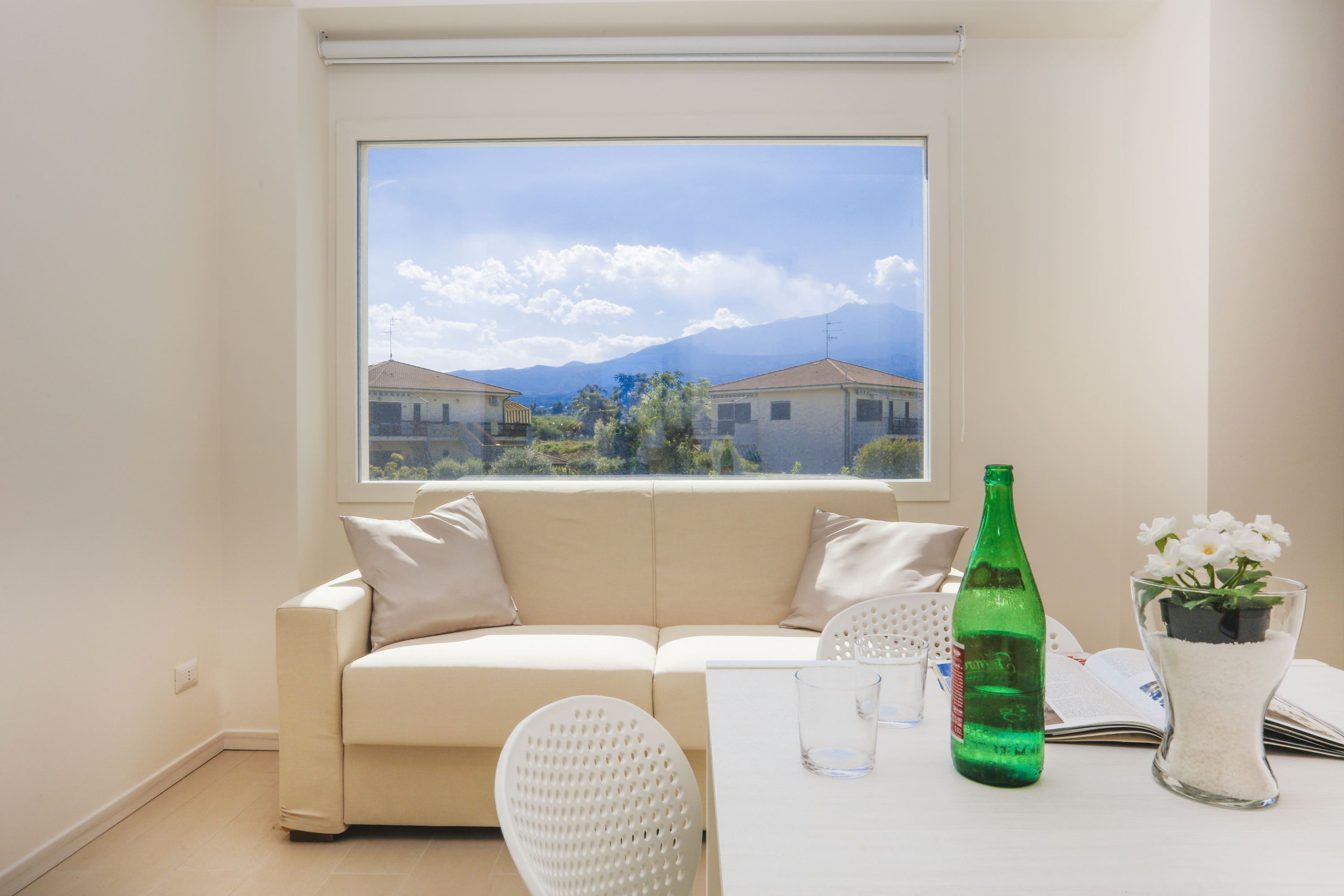 Una stanza con un divano beige, una sedia bianca, un tavolo con una bottiglia verde, un vaso di vetro con fiori e una finestra vista case e montagne.