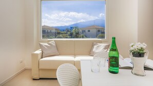 Una habitación con un sofá color beige, una silla blanca, una mesa con una botella verde, un florero de vidrio con flores y una ventana con vistas a casas y montañas.