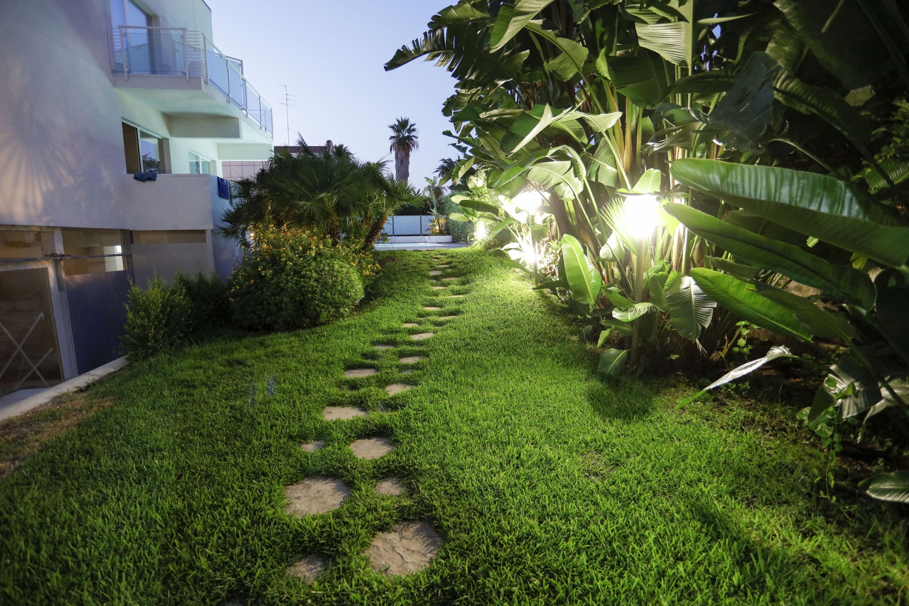 Un giardino curato con un vialetto in pietra, vegetazione rigogliosa e un edificio moderno con balconi.