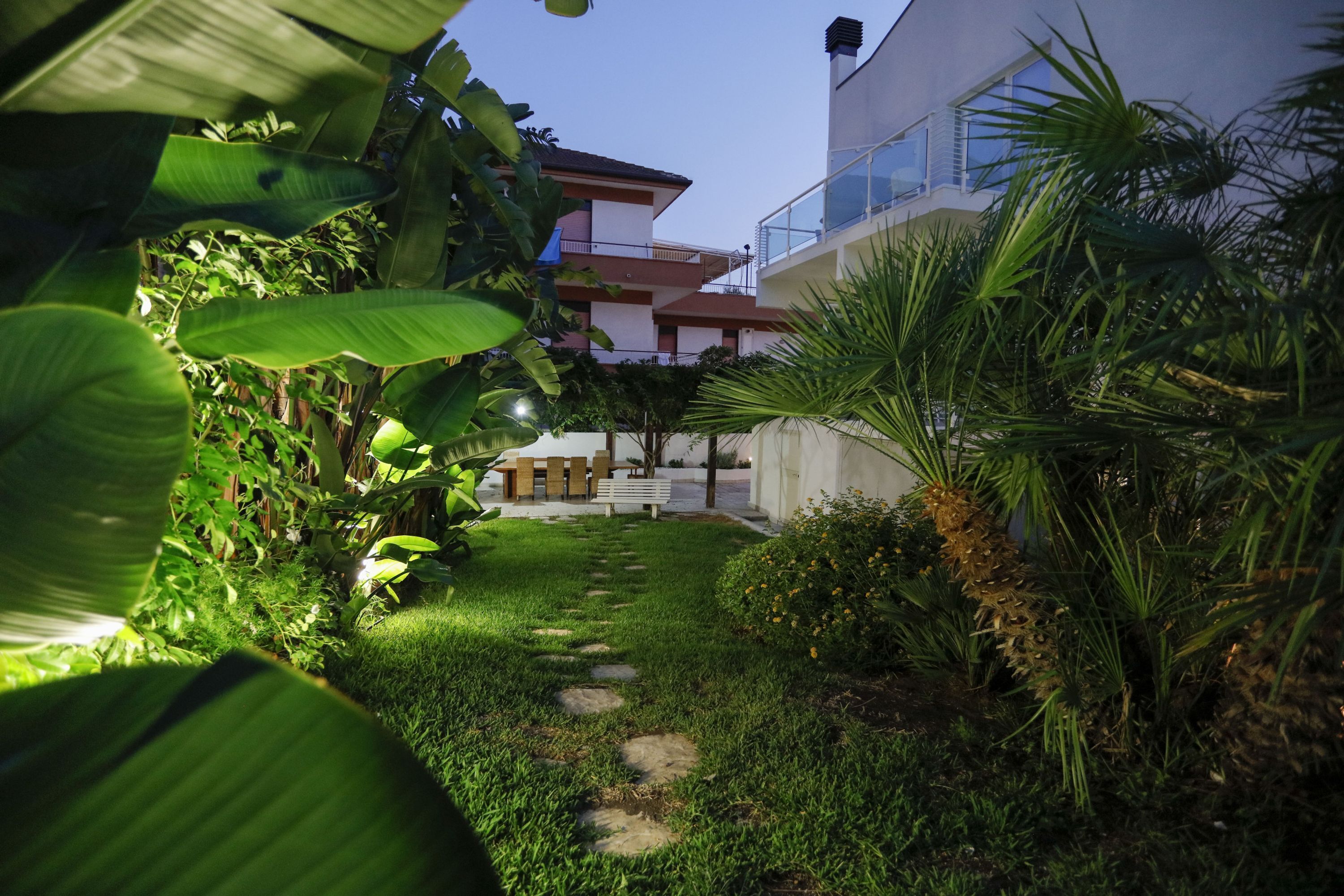 Un sendero en un jardín conduce a un edificio con balcón, rodeado de abundante vegetación y plantas tropicales.