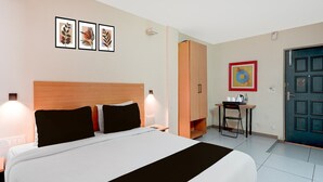 Hotellihuone, jossa on sänky, yöpöydällä olevat lamput, työpöytä tuolilla, vaatekaappi ja seinällä tauluja.